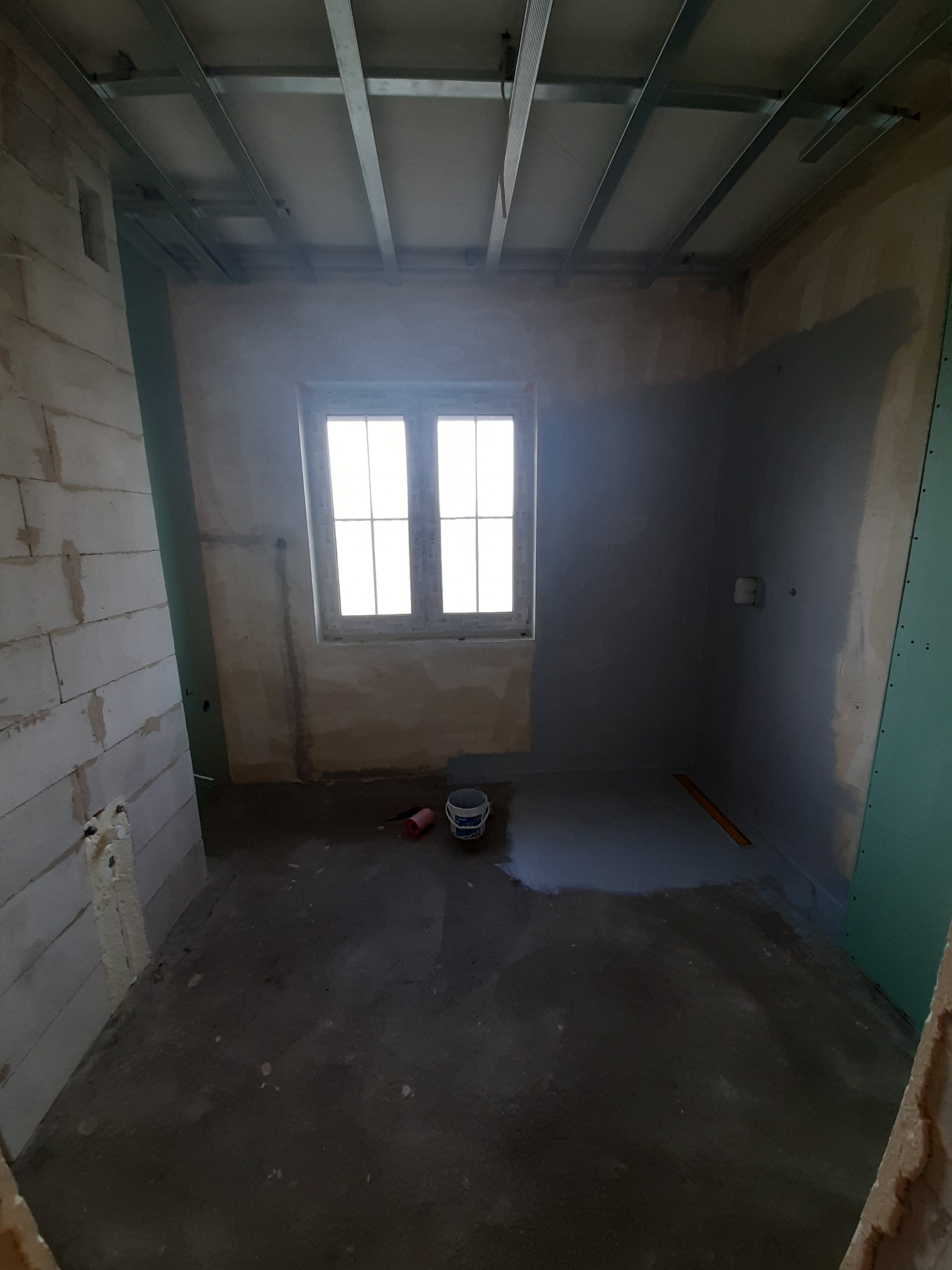 Wnętrze w trakcie remontu: ściany częściowo pokryte gładzią, sufit z konstrukcją metalową, okno z białymi ramami, na podłodze wiadro i poziomica.
