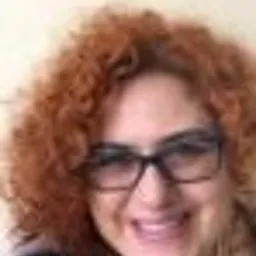 Portret kobiety z kręconymi rudymi włosami i okularami w czarnych oprawkach, uśmiechająca się delikatnie w jasnym wnętrzu.