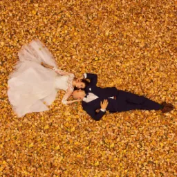 Para młoda leżąca na dywanie z jesiennych liści, widok z góry, on w granatowym garniturze, ona w białej sukni z koronkowymi rękawami.