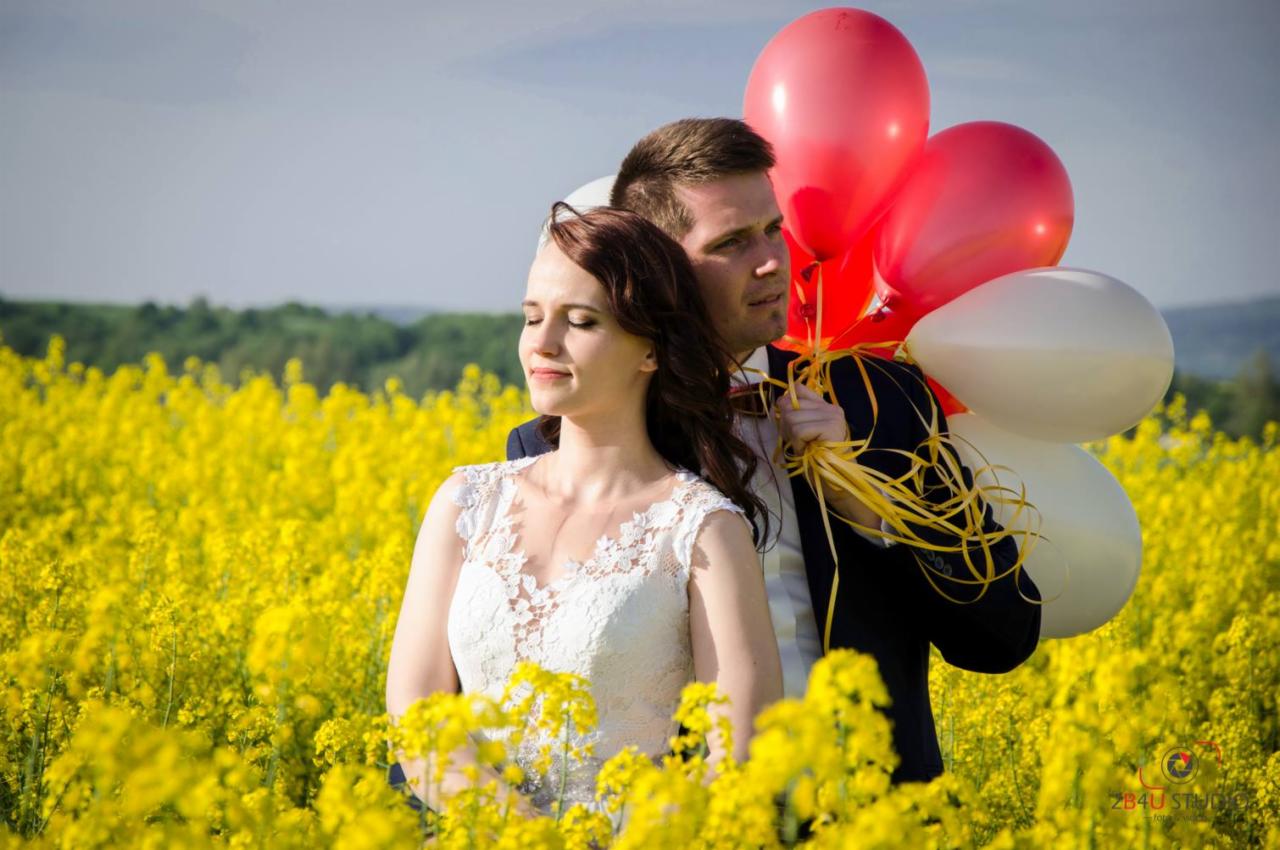 Para młoda w polu rzepaku, panna młoda w koronkowej sukni z zamkniętymi oczami, pan młody trzyma czerwone i białe balony.