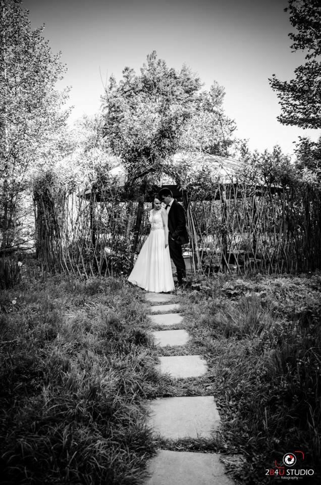 Czarno-biała fotografia pary młodej w ogrodzie, panna młoda w białej sukni, pan młody w czarnym garniturze, ścieżka z kamiennych płyt prowadzi do altany z gałęzi.