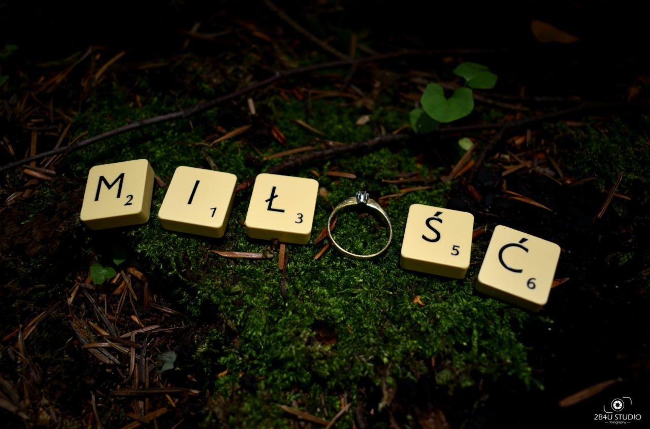 Pierścionek zaręczynowy umieszczony między literami 'MIŁOŚĆ' z gry Scrabble ułożonymi na mchu i igliwiu w leśnym otoczeniu, w tle widoczne gałązki