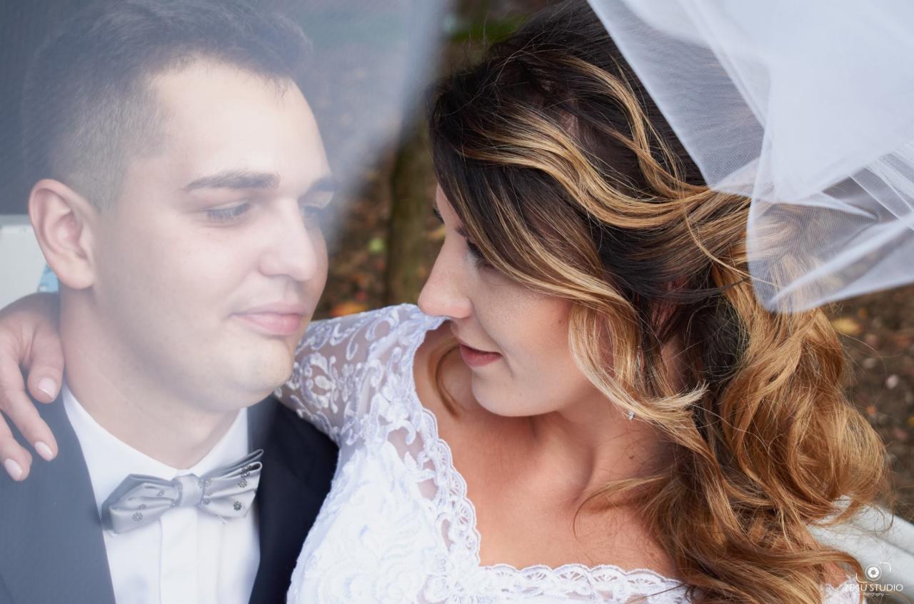 Panna młoda w koronkowej sukni i welonie, spogląda na pana młodego w ciemnym garniturze i srebrnej muszce, delikatne ujęcie przez przezroczystą zasłonę.