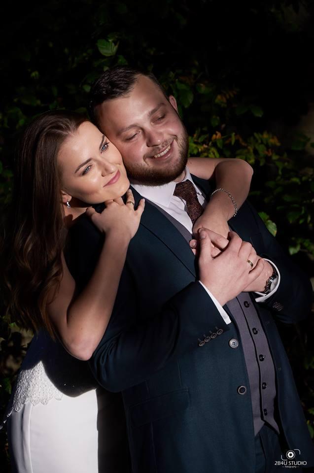 Uśmiechnięta para młoda w objęciach na tle ciemnej zieleni, panna młoda w sukni z koronkowym wykończeniem, pan młody w granatowym garniturze i bordowym krawacie.
