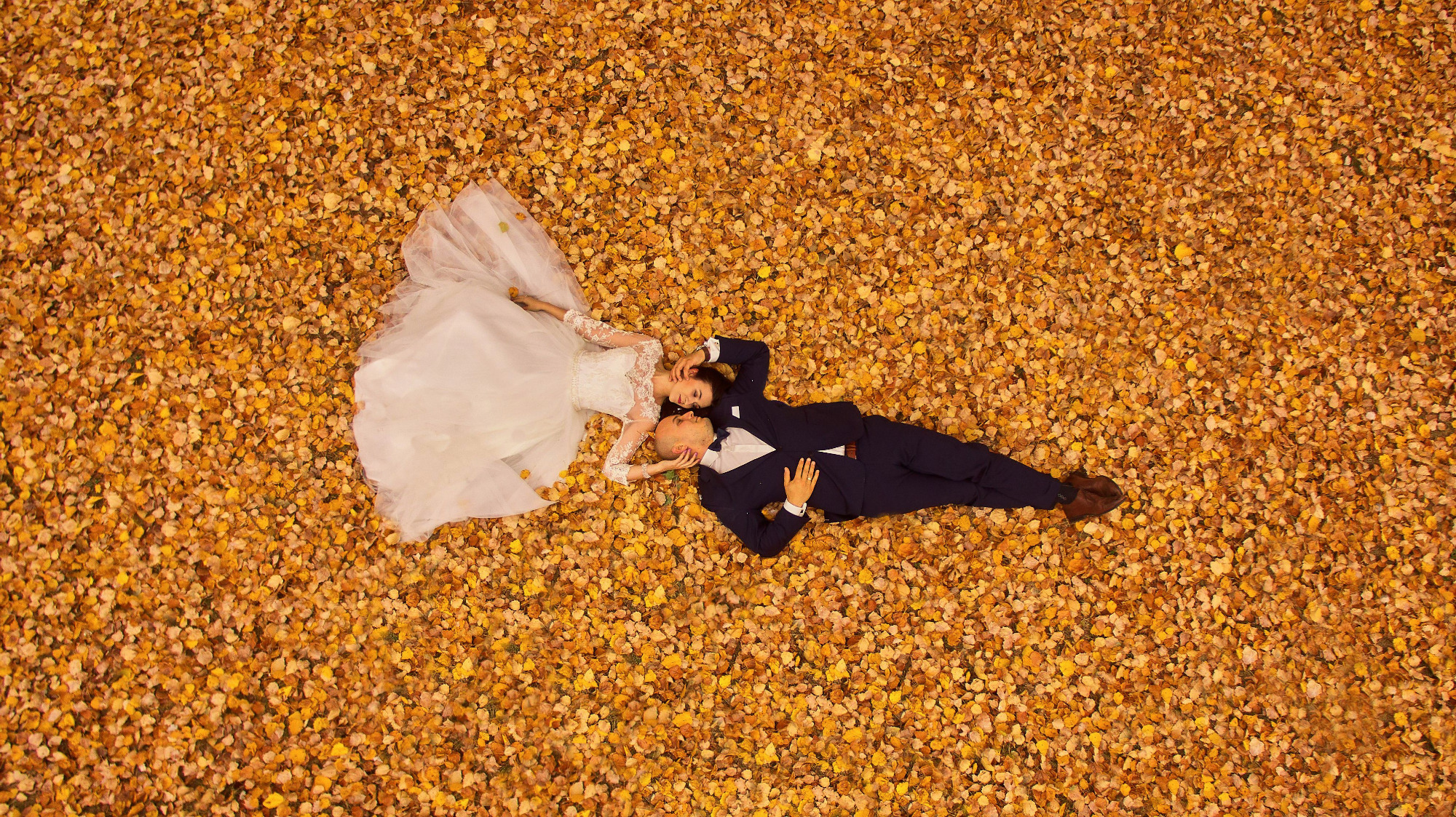 Para młoda leżąca na dywanie z jesiennych liści, widok z góry, on w granatowym garniturze, ona w białej sukni z koronkowymi rękawami.