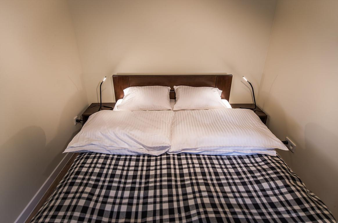 Dwuosobowe łóżko z drewnianym zagłówkiem, białą pościelą w prążki i czarno-białą kratą na narzucie, oświetlone lampkami nocnymi.