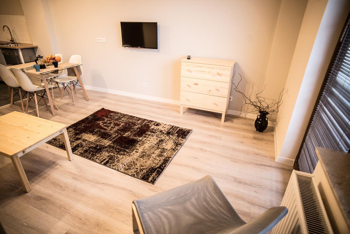 Jasne, nowocześnie urządzone wnętrze: drewniana podłoga, stół, krzesła, komoda, dywan w odcieniach brązu i beżu, telewizor na ścianie oraz dekoracyjna czarna waza z suchymi gałęziami.