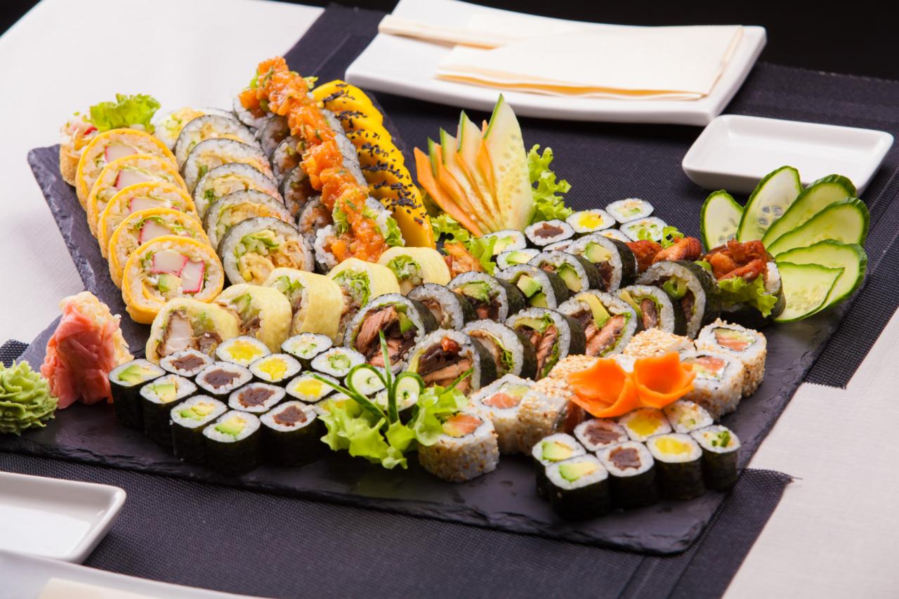 Bogaty zestaw sushi na czarnym kamiennym talerzu, z różnorodnymi rolkami, w tym z łososiem, awokado i warzywami, udekorowany rzeźbionymi ogórkami i marchewką, podany z imbirem i wasabi, na tle...
