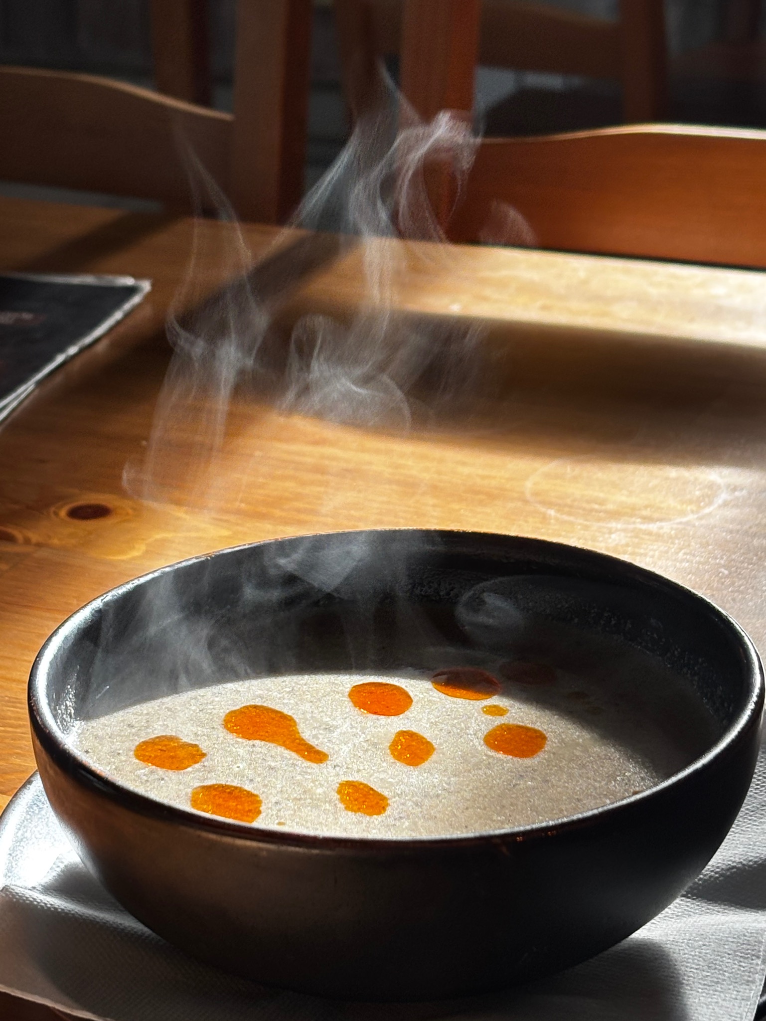 Parująca kremowa zupa w czarnej misce na drewnianym stole, z kroplami pomarańczowego oleju i delikatnym oświetleniem słonecznym.