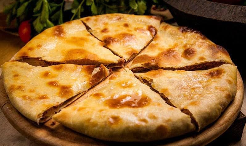 Osetyński placek z mięsem, pokrojony na trójkątne kawałki, leży na drewnianej desce. W tle zieleń i ciemny garnek.