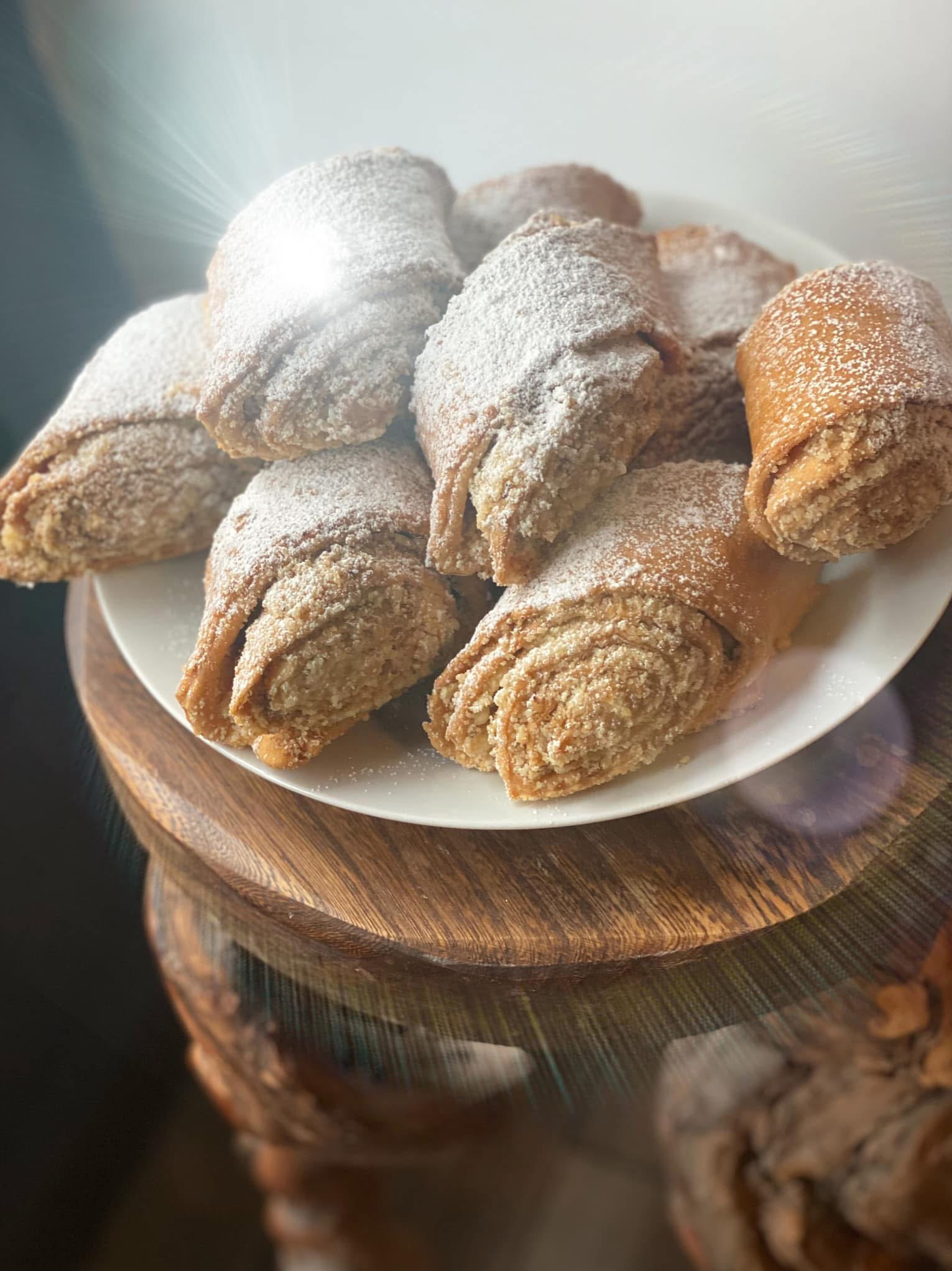 Kruche, złociste rogaliki posypane cukrem pudrem, ułożone na białym talerzu stojącym na drewnianym, rzeźbionym postumencie. Słodkie wypieki w zbliżeniu.