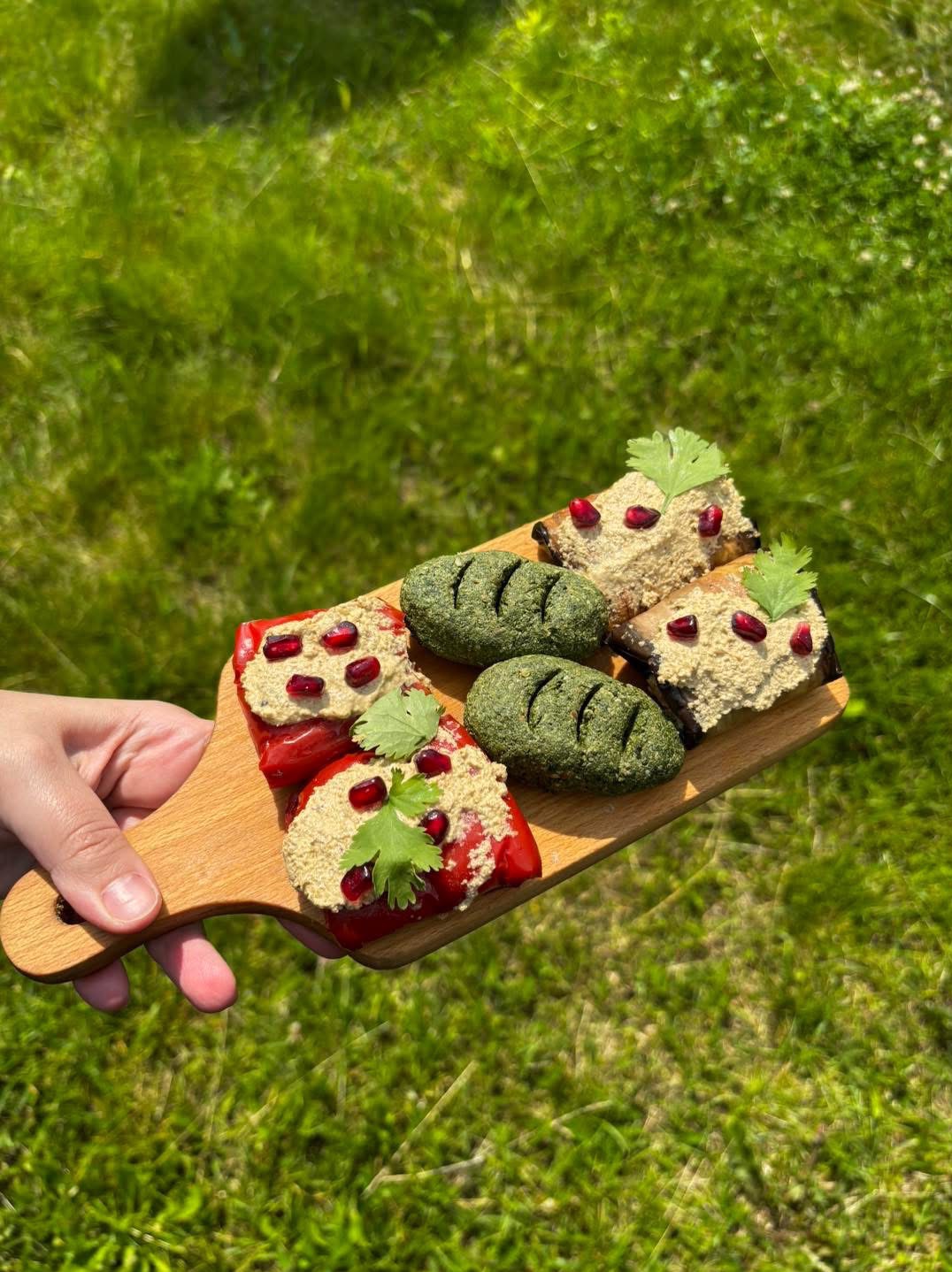 Ręka trzyma drewnianą deskę z gruzińskimi przekąskami: papryka i bakłażan z pastą orzechową, posypane granatem i zieloną kolendrą, na tle trawy.