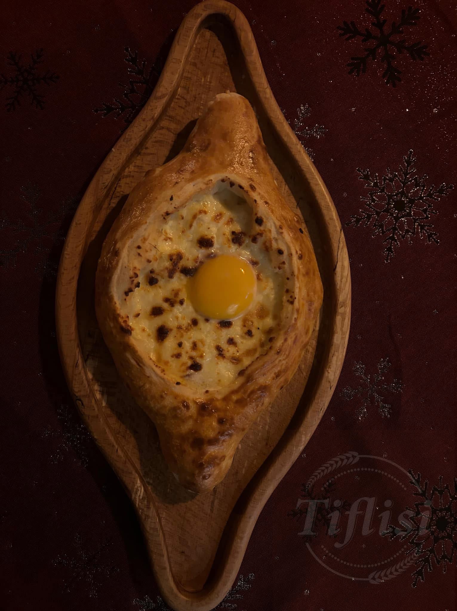 Adżarskie chaczapuri na drewnianej desce, z płynnym żółtkiem pośrodku, na tle bordowego obrusu w śnieżynki. Widoczne logo 'Tiflisi'.