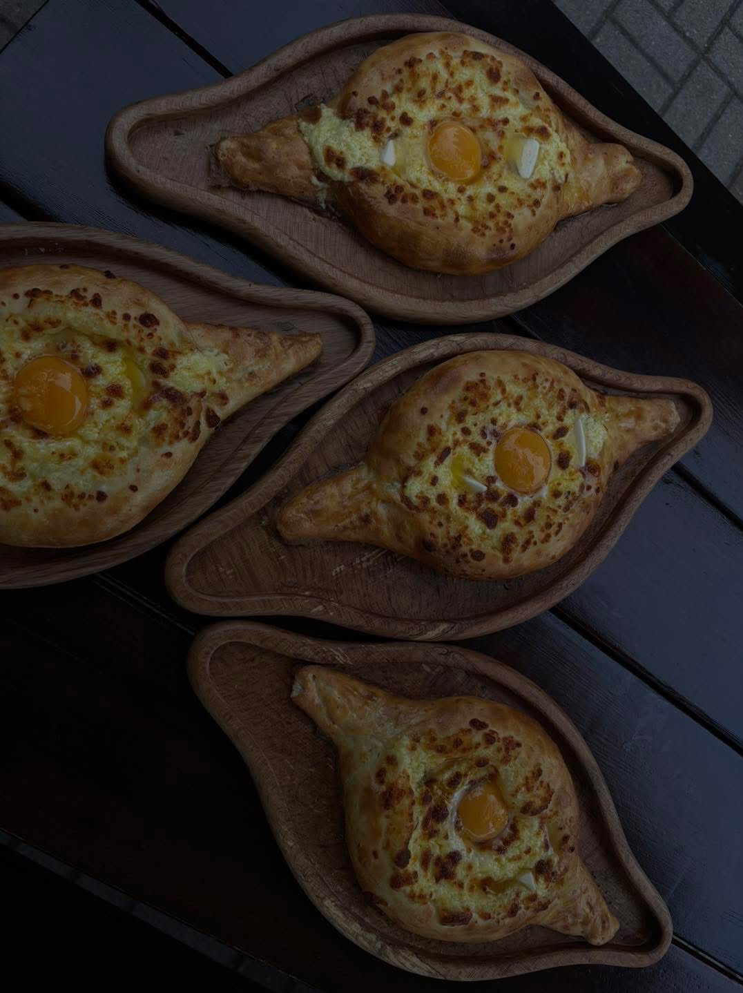 Cztery gruzińskie chaczapuri adżaruli z jajkiem na drewnianych tackach w kształcie liścia, ułożone na ciemnym blacie. Widok z góry.