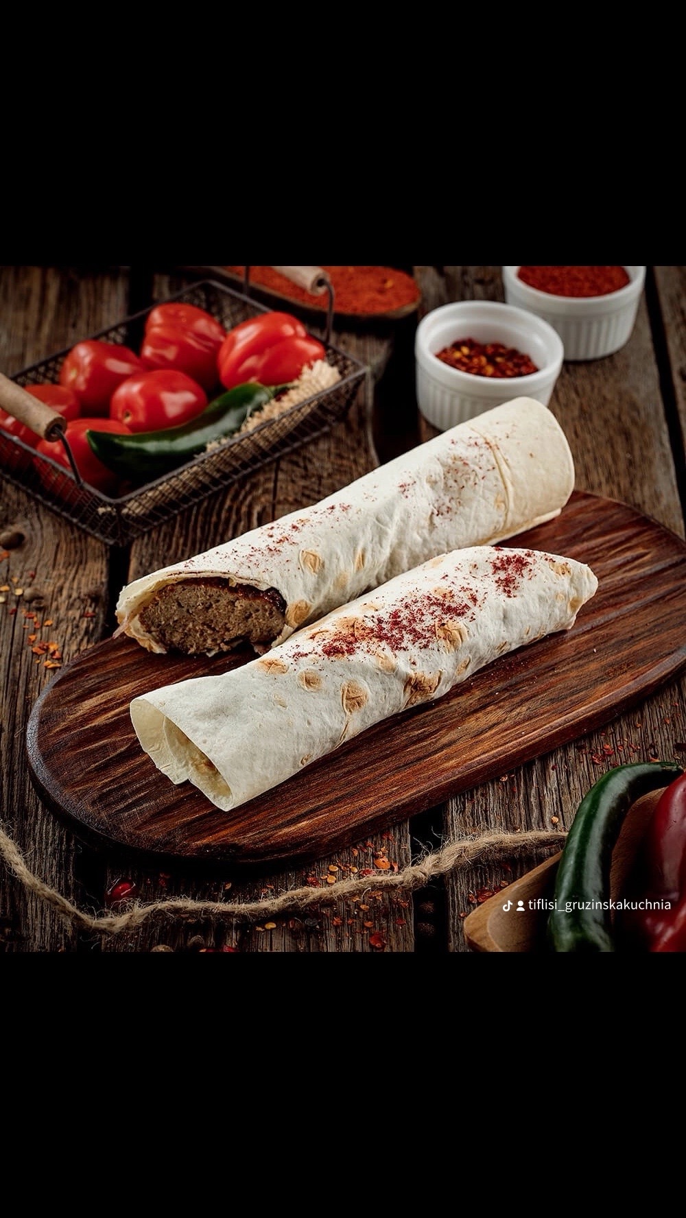 Dwa shawarmy na drewnianej desce, posypane przyprawami, z koszykiem pomidorów i papryczek w tle. Klimatyczne ujęcie potrawy z bliska.