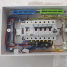 Rozdzielnica elektryczna w białej obudowie z widocznymi bezpiecznikami, przewodami w różnych kolorach i listwami zaciskowymi, zamontowana na nierównej, białej ścianie.