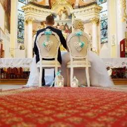Para młoda siedzi na krzesłach z dekoracjami w kształcie serc w kościele, widok z tyłu, czerwony dywan prowadzi do ołtarza.