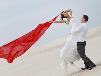 Uśmiechnięta panna młoda w białej sukni, niesiona przez pana młodego na piaszczystej wydmie, trzymająca powiewającą czerwoną tkaninę.