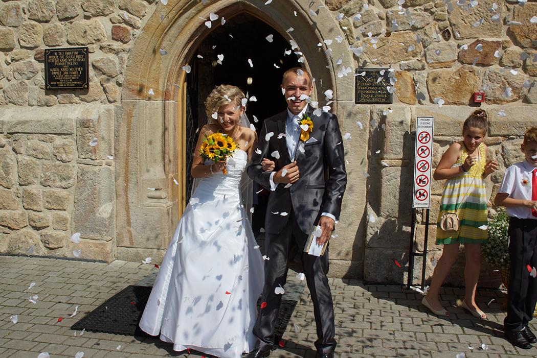 Uśmiechnięta para młoda wychodzi z kamiennego kościoła, obsypywana płatkami kwiatów; bukiet słoneczników w dłoni panny młodej, ciemny garnitur pana młodego z żółtą butonierką, w tle osoby obserwujące.