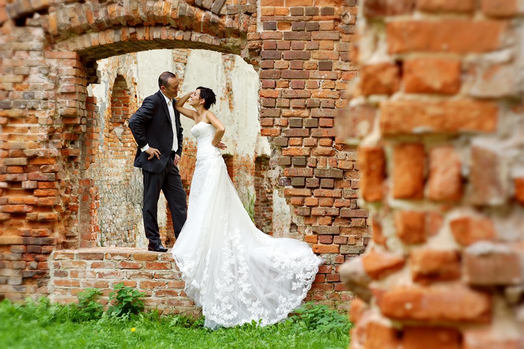 Para młoda pozuje na tle ceglanego, zrujnowanego muru z prześwitem, on w czarnym garniturze, ona w białej sukni z koronkowymi aplikacjami, dotyka jego twarzy.