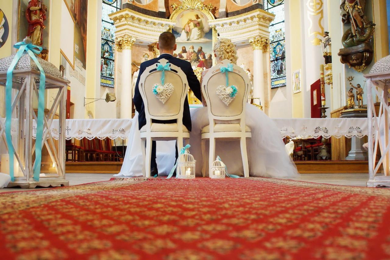 Para młoda siedzi na krzesłach z dekoracjami w kształcie serc w kościele, widok z tyłu, czerwony dywan prowadzi do ołtarza.