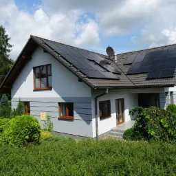 Dwukondygnacyjny dom jednorodzinny z szarą elewacją, brązowymi oknami i panelami słonecznymi na dachu, otoczony zielenią w słoneczny dzień.