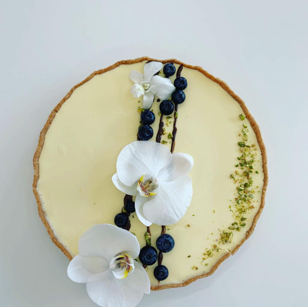 Tarta z kremem, dekorowana białymi orchideami, borówkami i posiekanymi pistacjami na białym tle.