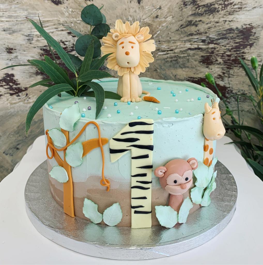 Tort urodzinowy dla roczniaka z motywem safari: lew, żyrafa, małpka i cyfra '1' w paski zebry, otoczone zielonymi liśćmi i gałązkami.