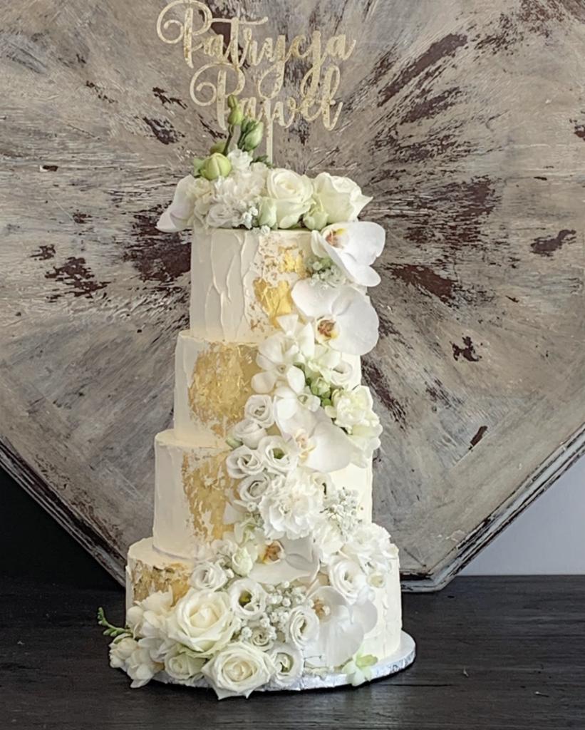 Czteropiętrowy tort weselny z białym kremem, płatkami złota, kaskadą białych kwiatów i topperem z imionami 'Patrycja i Paweł' na tle dekoracyjnej ściany.