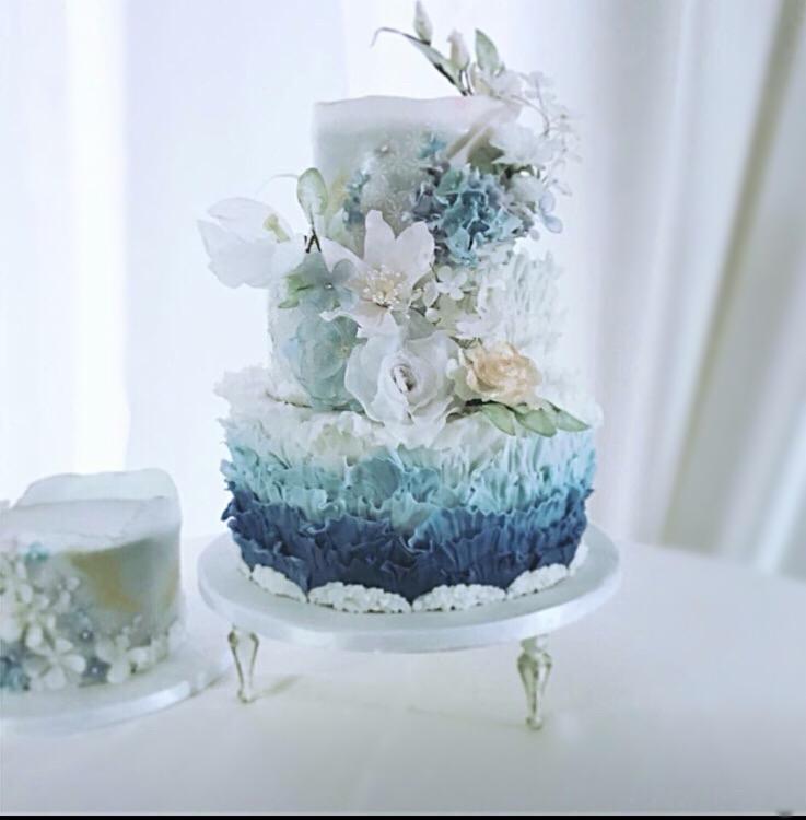 Trzypiętrowy tort ombre w odcieniach bieli i błękitu, udekorowany białymi i niebieskimi kwiatami z lukru, na srebrnym stojaku z nóżkami, obok mniejszy tort z podobnymi dekoracjami.