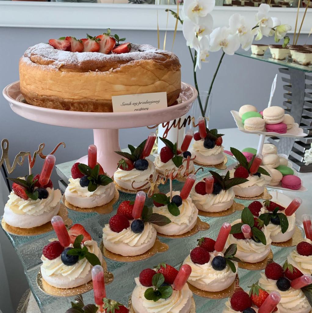 Sernik posypany cukrem pudrem i udekorowany truskawkami na różowej paterze, obok mini bezy z owocami i pipetą z sosem oraz makaroniki na szklanym stole.