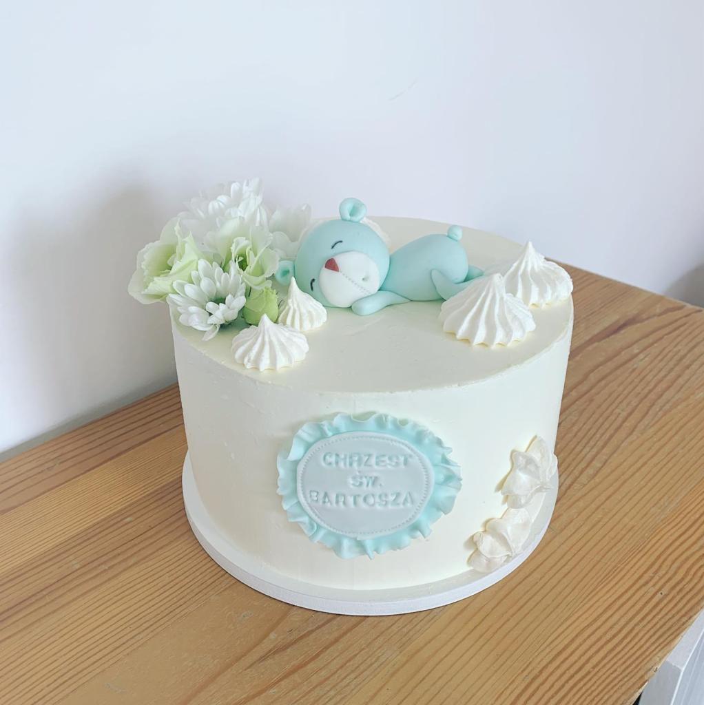 Tort okolicznościowy z białym kremem, dekorowany niebieską figurką misia śpiącego na wierzchu, białymi bezami i kwiatami, z napisem 'Chrzest Św. Bartosza' w błękitnej ramce.