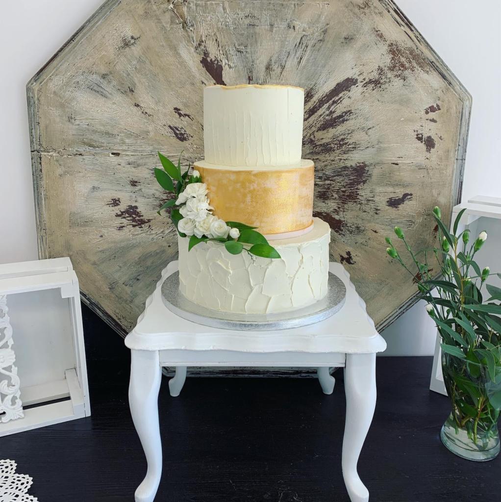 Trzypiętrowy tort weselny z kremowym lukrem, złotą środkową warstwą i dekoracją z białych kwiatów, ustawiony na białym stoliku na tle rustykalnej drewnianej dekoracji ściennej.