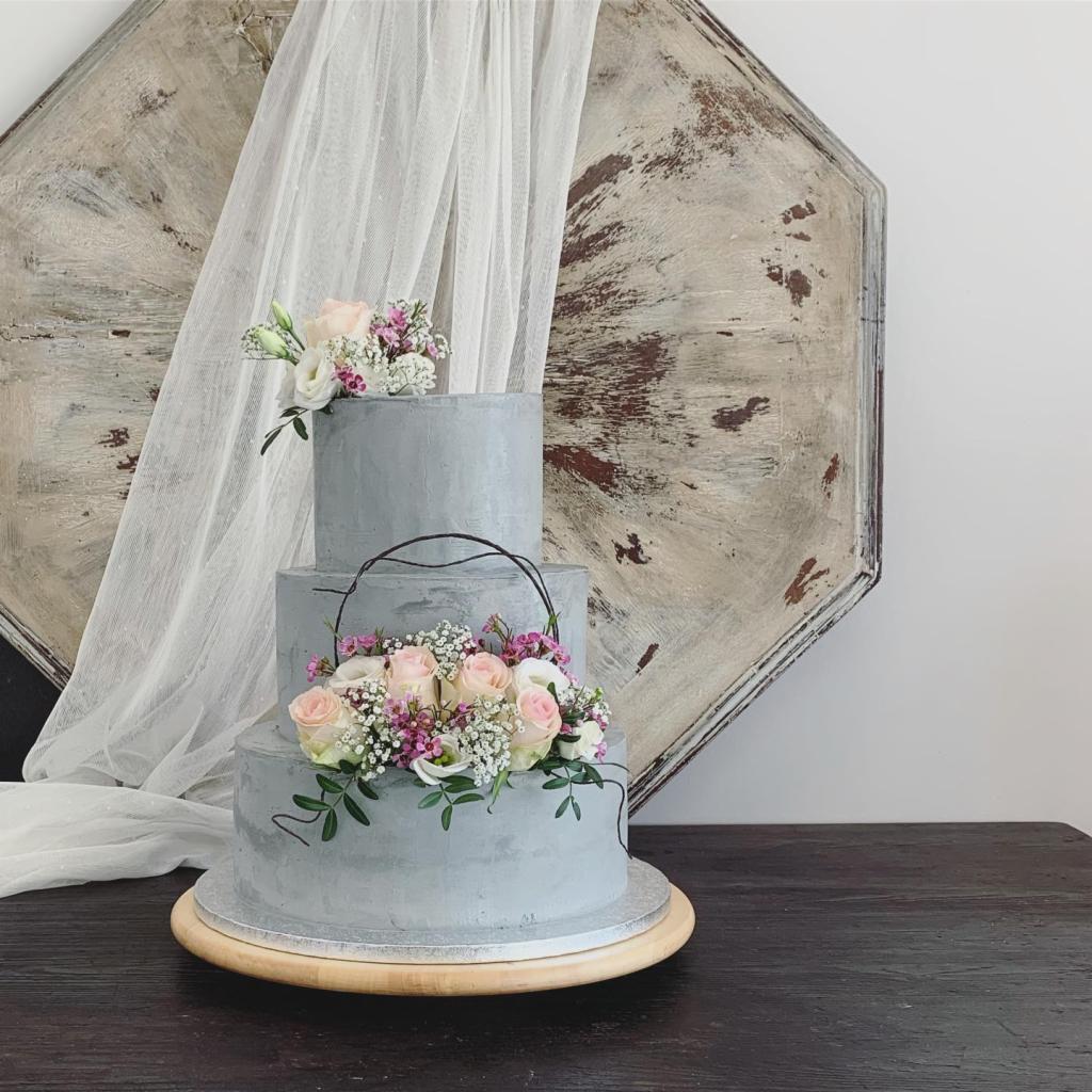 Trzypiętrowy tort weselny w odcieniach szarości, udekorowany kompozycjami z różowych i białych róż oraz drobnych kwiatów, na drewnianym podkładzie, z dekoracyjną tkaniną i drewnianą ozdobą w tle.