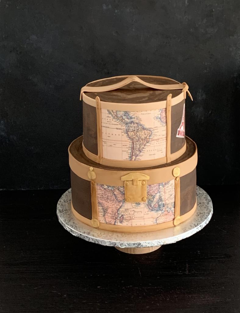 Dwupoziomowy tort w kształcie walizki podróżnej z mapami świata i wieżą Eiffla, na srebrnej paterze.