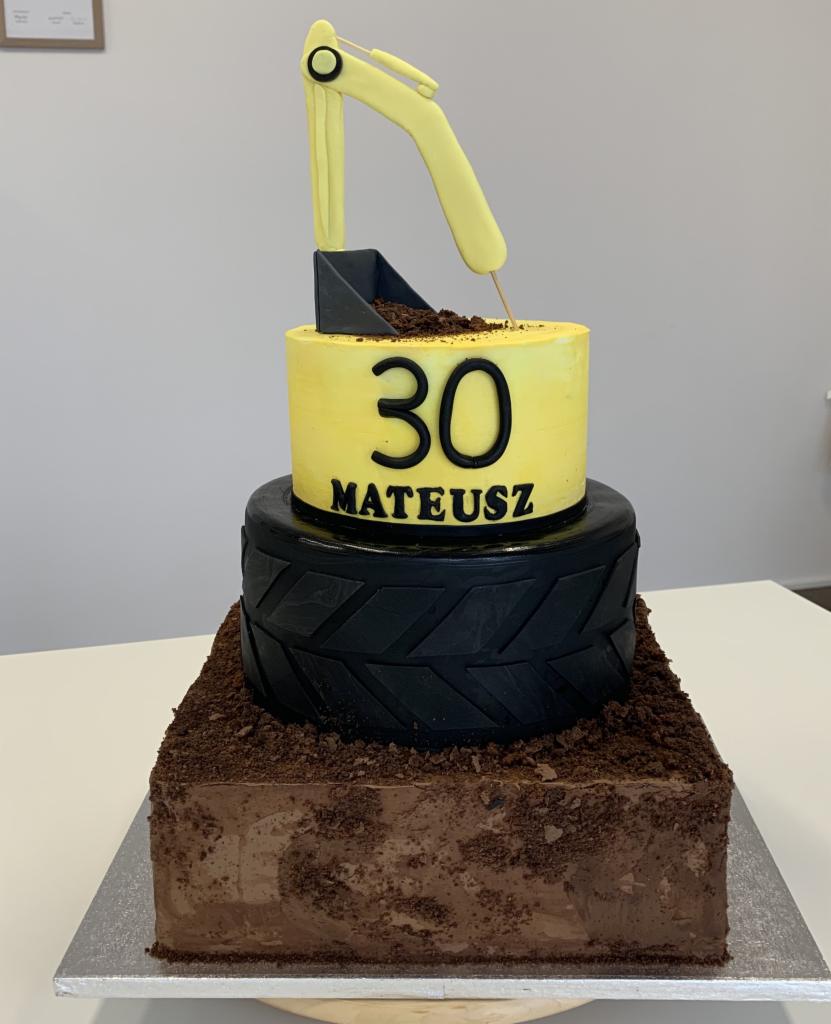 Tort urodzinowy w kształcie koparki, z żółtym korpusem z napisem '30 Mateusz', czarnym kołem jako środkowa warstwa i czekoladową podstawą, całość na srebrnej tacy.