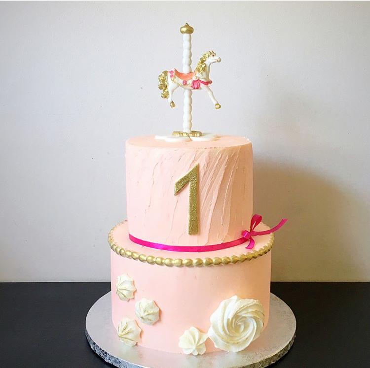 Dwupoziomowy tort w odcieniach różu z dekoracją w postaci karuzeli z konikiem i złotą cyfrą '1', ozdobiony różową wstążką i bezami.