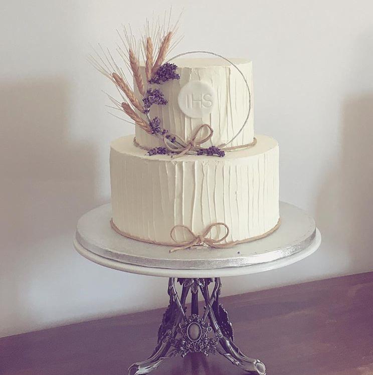 Dwupoziomowy tort komunijny z kremowym lukrem o fakturze pionowych pasków, ozdobiony suszonymi kwiatami lawendy, kłosami pszenicy i symbolem IHS na górnym poziomie, umieszczony na srebrnym...