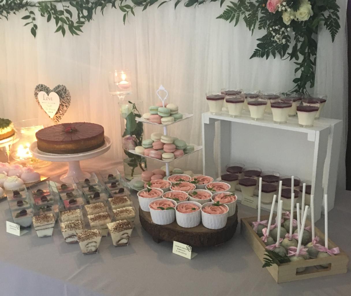 Dekoracyjny stół z różnorodnymi deserami: tort czekoladowy z truskawką, mini serniczki, desery w kubeczkach, makaroniki, babeczki z różowym kremem i cake pops, ustawione na różnych poziomach...