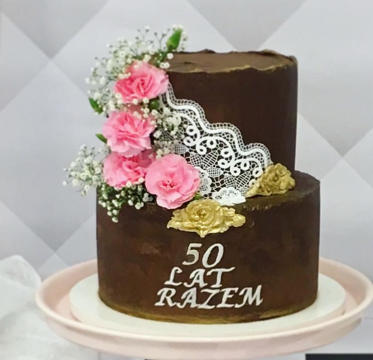 Dwupoziomowy tort czekoladowy z napisem '50 lat razem', ozdobiony różowymi goździkami, gipsówką, koronkową dekoracją i złotymi akcentami, umieszczony na różowym talerzu.