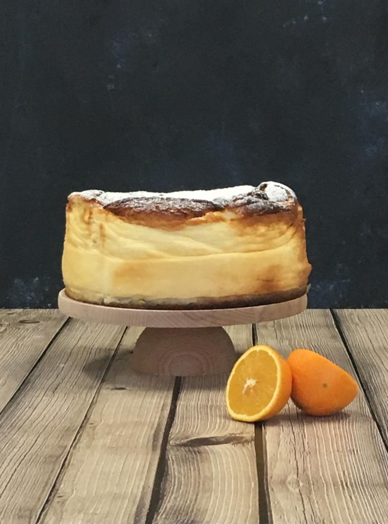 Wypiekany sernik baskijski posypany cukrem pudrem na drewnianej paterze, obok dwie pomarańcze na tle ciemnego tła i drewnianego stołu.