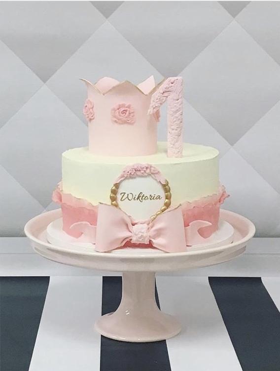 Dwupoziomowy tort urodzinowy w odcieniach różu i kremu, ozdobiony koroną, cyfrą '1' i dekoracyjnym napisem 'Wiktoria', umieszczony na różowej paterze na tle czarno-białych pasów.