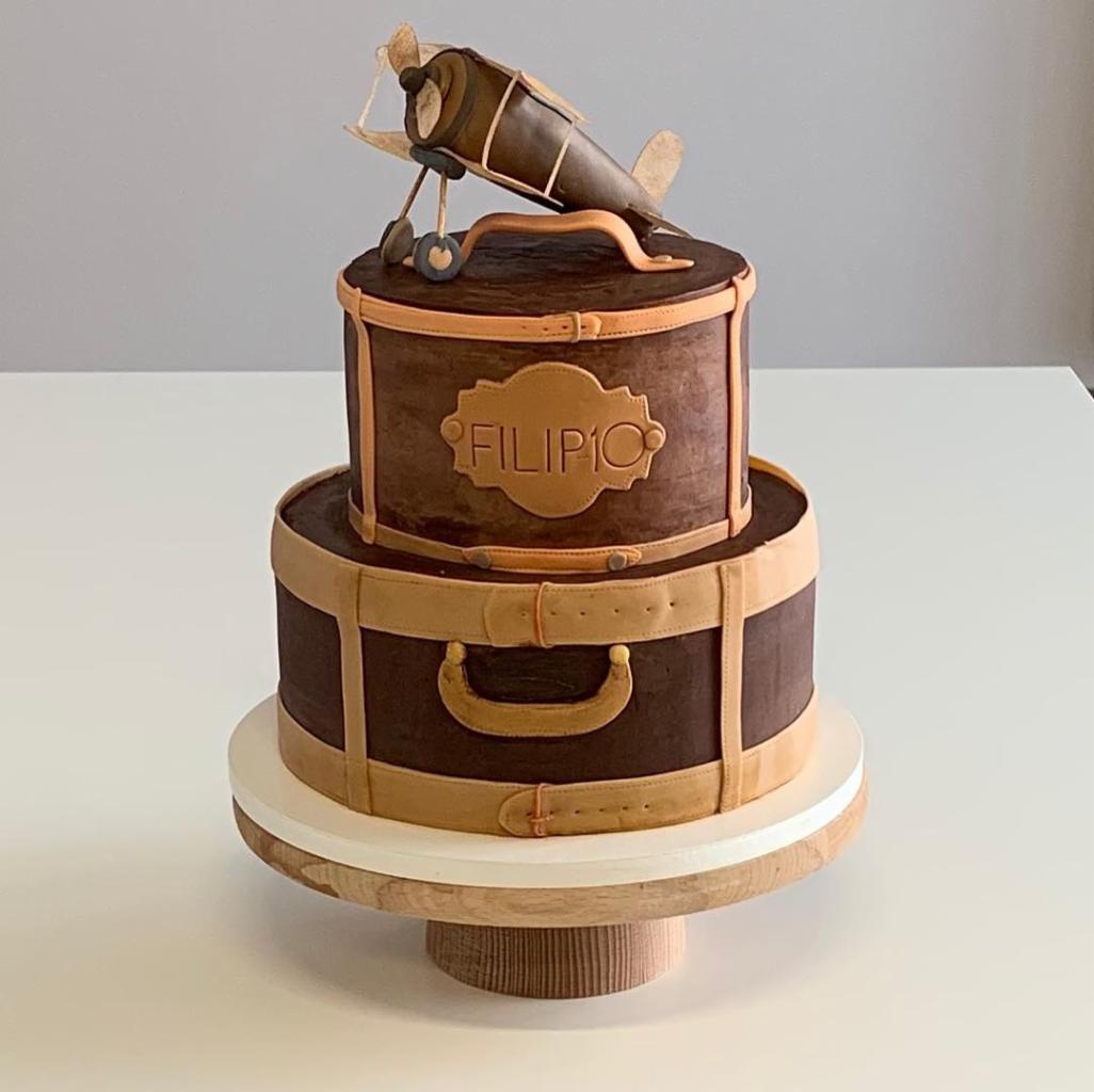 Dwupoziomowy tort w kształcie walizek podróżnych, ozdobiony modelem samolotu z brązowego lukru, umieszczony na drewnianym stojaku.