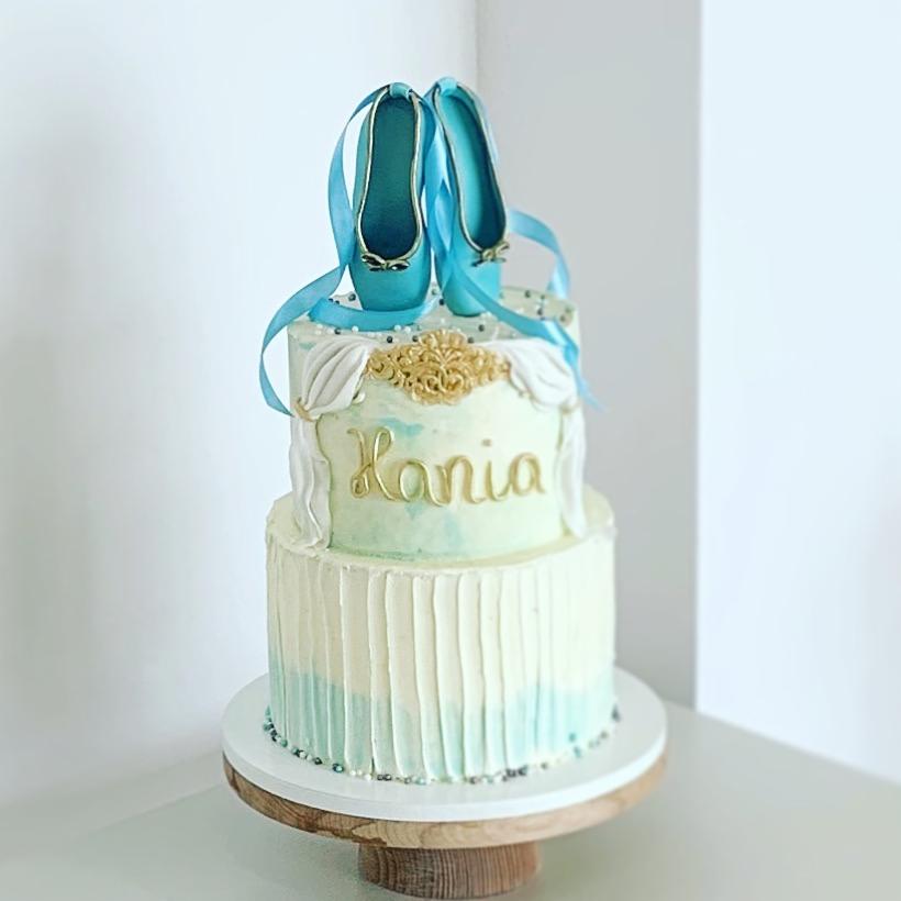 Dwupiętrowy tort zdobiony niebieskimi baletkami z wstążkami, złotym napisem 'Hania' i dekoracjami z kremu, umieszczony na drewnianej podstawie.