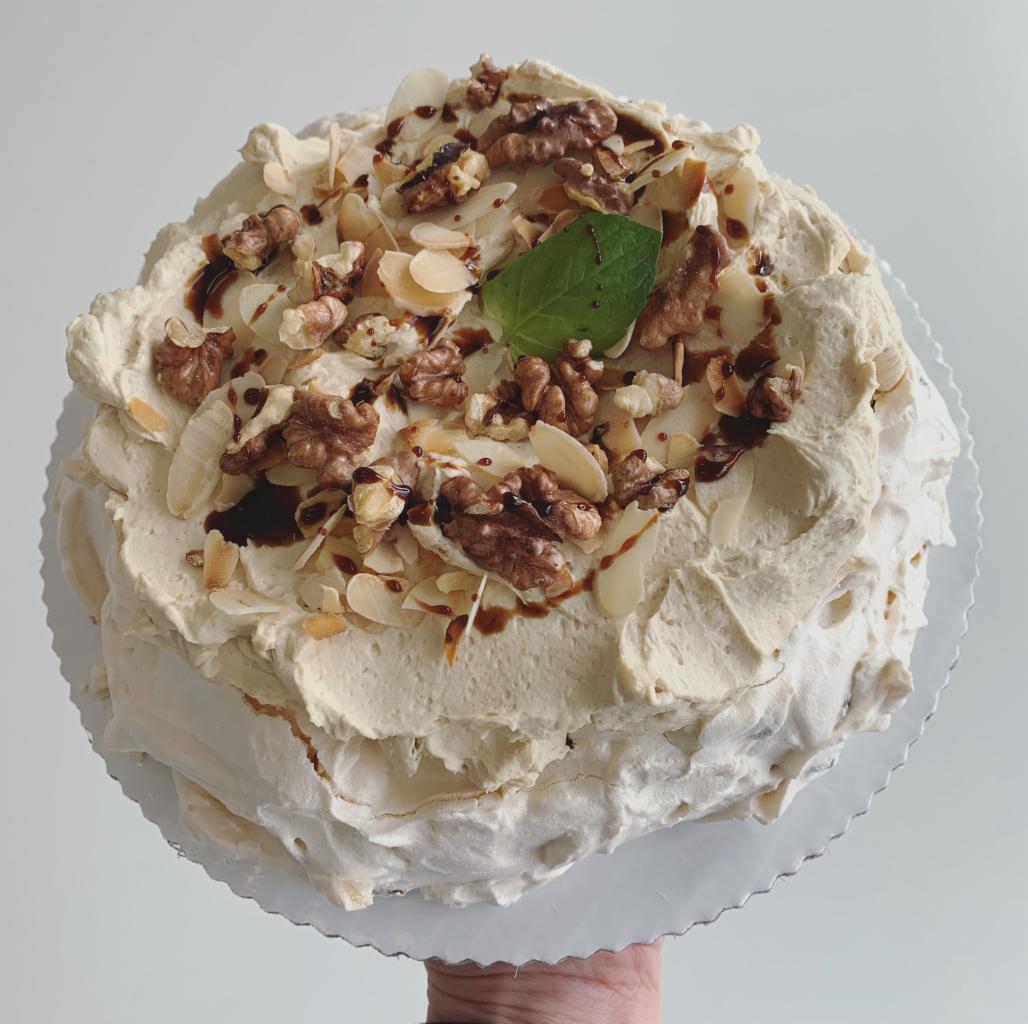 Beżowy tort bezowy z orzechami włoskimi, płatkami migdałów, karmelem i listkiem mięty, na srebrnej podkładce, trzymany w dłoni.