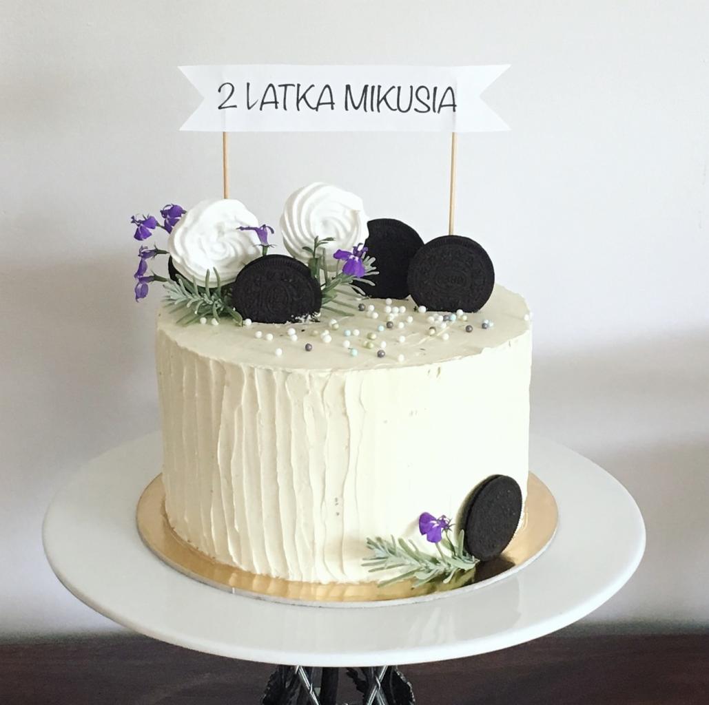 Tort urodzinowy dla dwulatka Mikusia, z kremowym lukrem, dekorowany Oreo, bezami, fioletowymi kwiatkami i posypką z perełek, umieszczony na białym paterze.