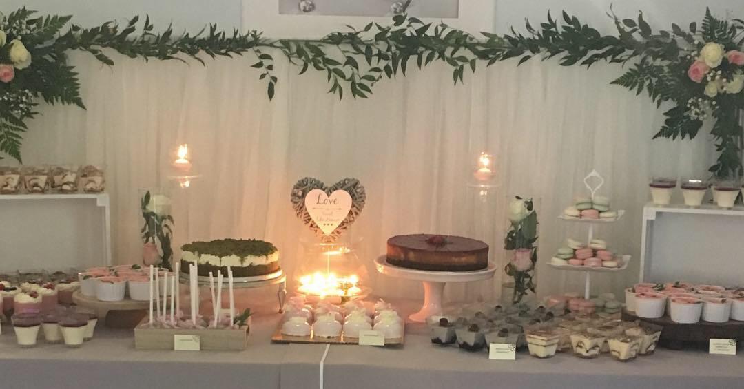 Stół z deserami na przyjęciu: torty, babeczki, makaroniki, tiramisu w małych pojemnikach, dekoracje kwiatowe i świece, serce z napisem 'Love'.
