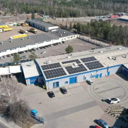 Widok z góry na budynek przemysłowy z zainstalowanymi panelami słonecznymi na dachu, na parkingu zaparkowane samochody, w tle inne budynki i drzewa.
