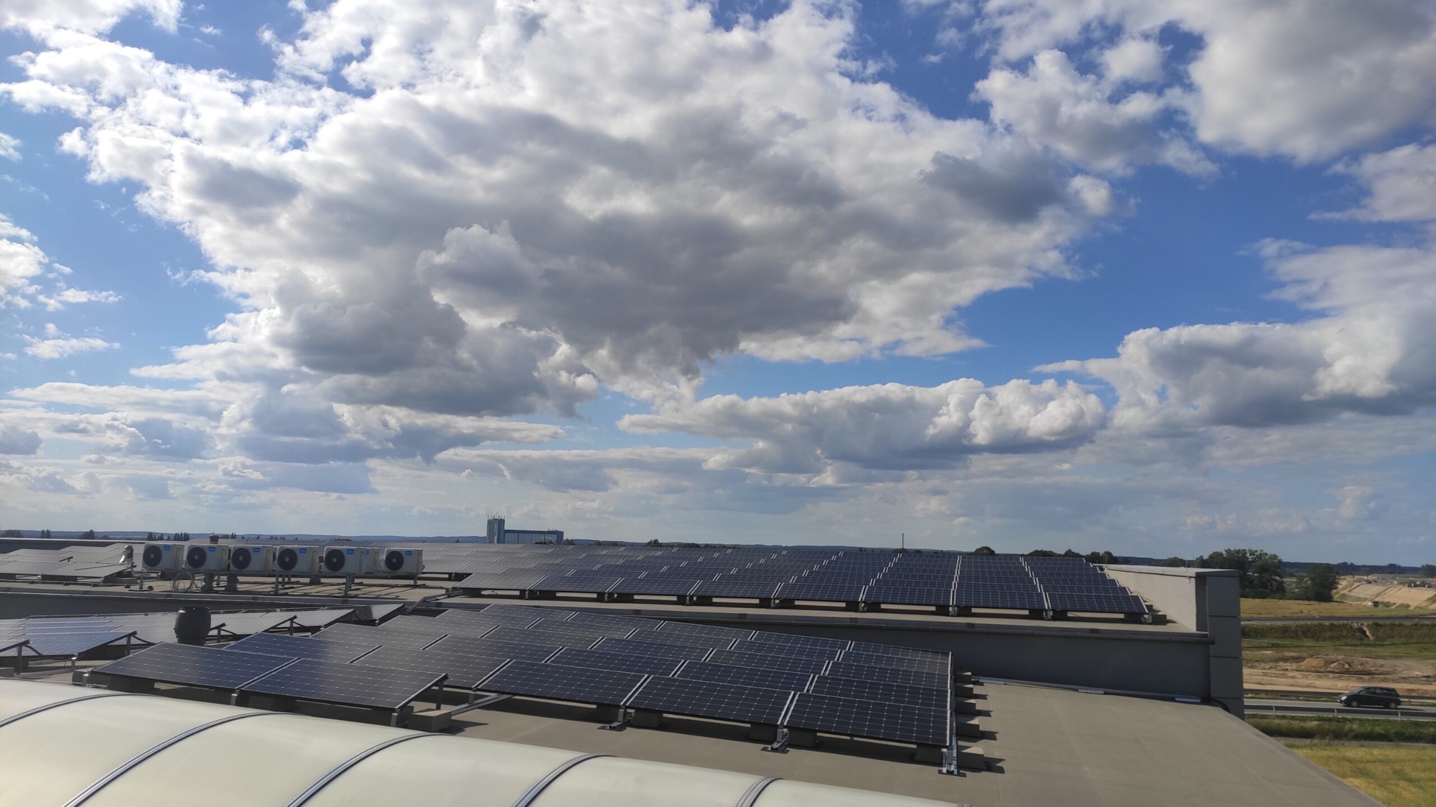 Rząd paneli słonecznych na płaskim dachu budynku przemysłowego, z rzędem klimatyzatorów w tle i błękitnym niebem z chmurami powyżej.