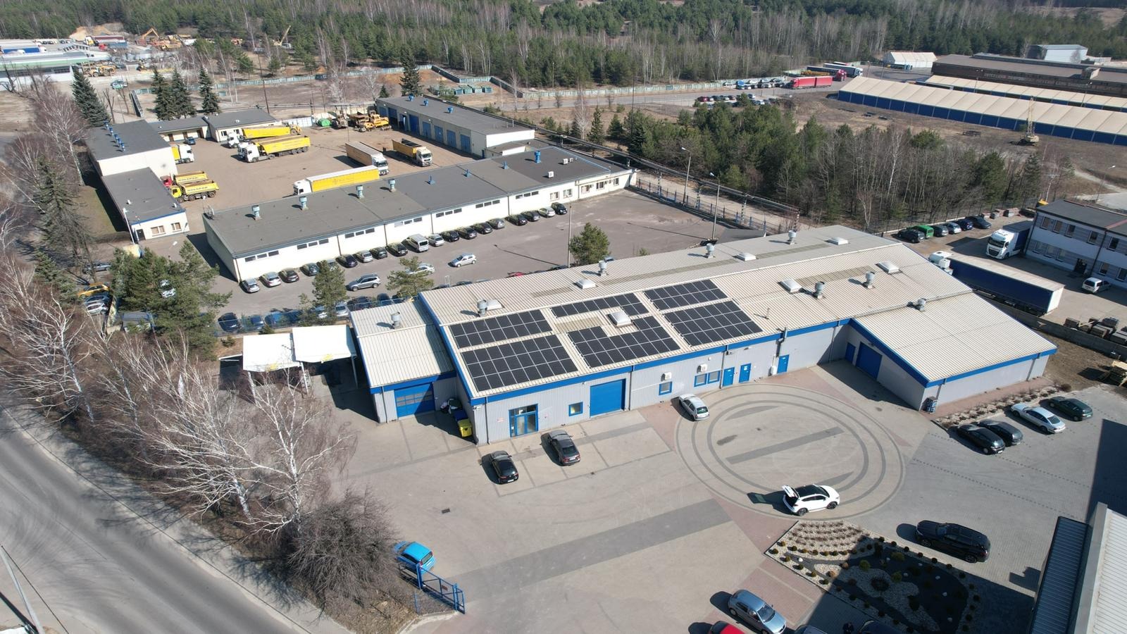 Widok z góry na budynek przemysłowy z zainstalowanymi panelami słonecznymi na dachu, na parkingu zaparkowane samochody, w tle inne budynki i drzewa.